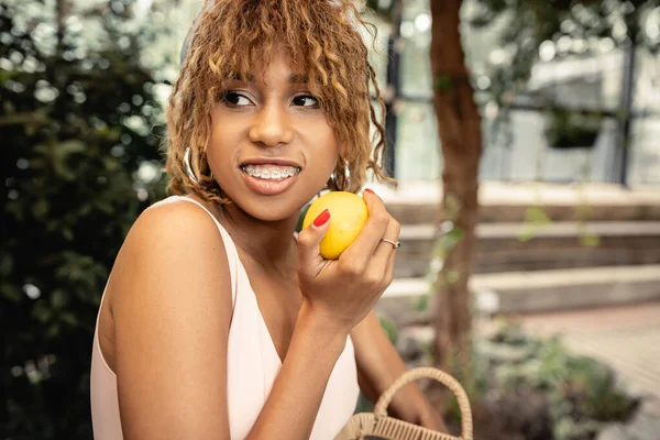 Porträt einer lächelnden jungen afrikanisch-amerikanischen Frau mit Zahnspange, die frische Zitrone und Korb hält, während sie im verschwommenen Gewächshaus im Hintergrund wegschaut, stilvolle Dame, die Mode und Natur vereint — Stockfoto