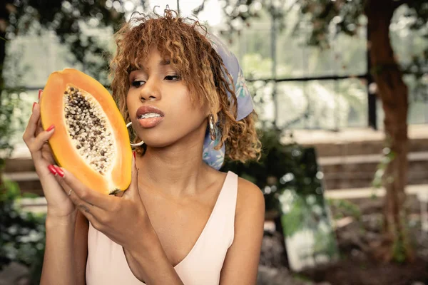 Portrait de jeune femme afro-américaine à la mode en foulard tenant papaye fraîche tout en passant du temps dans le centre de jardin flou, dame élégante mêlant mode et nature, concept d'été — Photo de stock