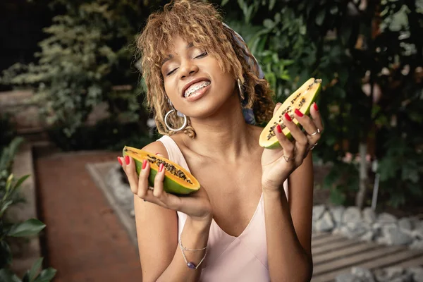 Smiling young african american woman with braces waring summer outfit and closing eyes while holding fresh papaya in blurred indoor garden, fashion-forward lady inspired by tropical plants — Stock Photo