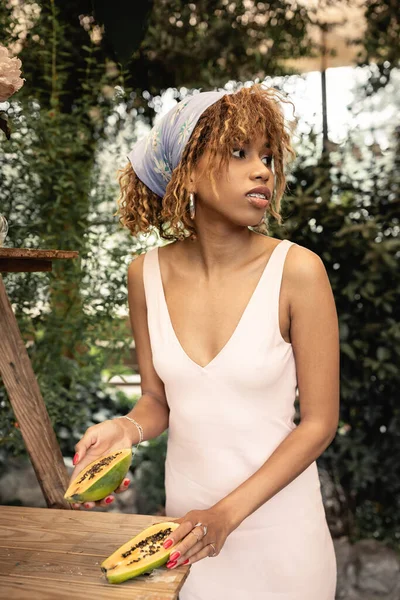 Joven mujer afroamericana en elegante pañuelo para la cabeza y vestido de verano sosteniendo papaya fresca y mirando hacia otro lado en invernadero borroso, señora de la moda hacia adelante inspirada en las plantas tropicales, concepto de verano - foto de stock