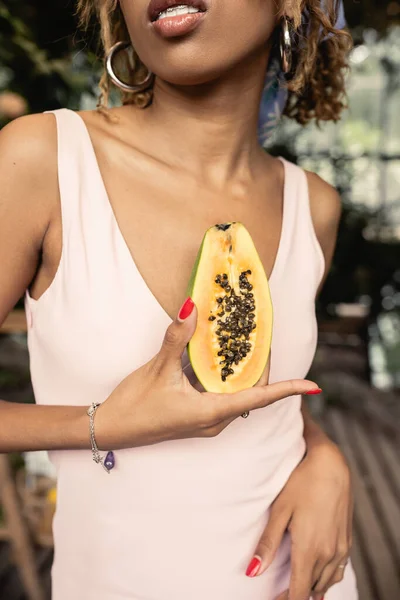 Vista recortada de la elegante joven afroamericana con tirantes que usan vestido de verano mientras sostiene papaya madura cortada en el centro del jardín borroso, dama de moda hacia adelante inspirada en las plantas tropicales - foto de stock