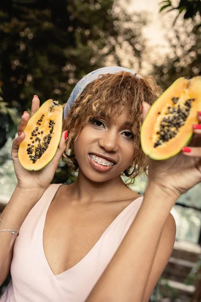 Joven y alegre mujer afroamericana con tirantes con vestido de verano y pañuelo en la cabeza mientras sostiene la papaya cortada y mira a la cámara en naranjería, dama de moda inspirada en las plantas tropicales - foto de stock