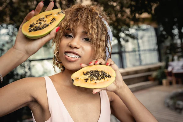 Portrait de jeune femme afro-américaine positive avec des bretelles en tenue d'été et tenant la papaye fraîche debout dans l'orangerie, femme à la mode entourée de luxuriance tropicale — Photo de stock