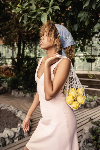 Vista lateral de la joven mujer afroamericana de moda en pañuelo para la cabeza y vestido de verano sosteniendo bolsa de puré con limones frescos mientras está de pie en el centro del jardín borroso, mujer de moda rodeada de exuberancia tropical - foto de stock