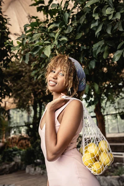 Portrait de femme afro-américaine souriante avec bretelles en robe d'été tenant un sac en filet avec des citrons frais et regardant la caméra dans le jardin intérieur, dame élégante appréciant l'atmosphère tropicale — Photo de stock