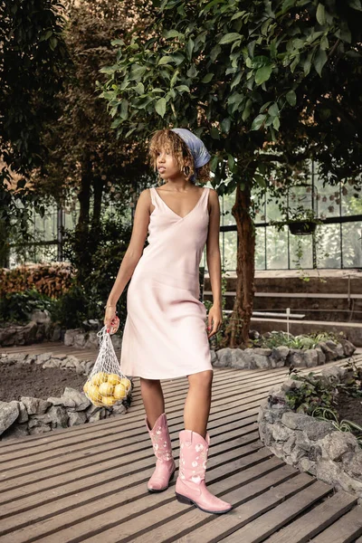 Mulher americana africana jovem na moda em vestido de verão segurando limões frescos em saco de malha e de pé no jardim interior borrado no fundo, senhora elegante desfrutando de atmosfera tropical — Fotografia de Stock