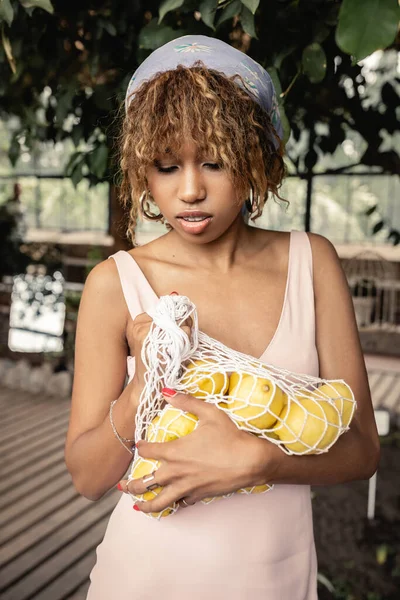 Retrato de una joven afroamericana con pañuelo en la cabeza y vestido de verano sosteniendo bolsa de malla con jugosos limones y de pie en un borroso naranjero de fondo, elegante dama disfrutando de la atmósfera tropical - foto de stock