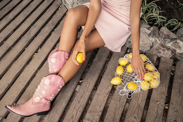 Top view of trendy young african american woman in summer dress and boots holding fresh lemon near mesh bag and sitting in garden center, chic woman in tropical garden, summer concept — Stock Photo