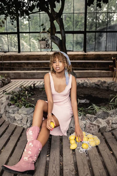 Full length of young frican american woman in summer outfit holding fresh lemon near mesh bag and looking at camera while sitting in garden center, chic woman in tropical garden, summer concept — Stock Photo
