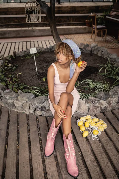 Mujer afroamericana joven de moda en traje de verano cruzando piernas y sosteniendo limón fresco mientras está sentado cerca de la bolsa de malla en el jardín interior, mujer elegante en el jardín tropical, concepto de verano - foto de stock