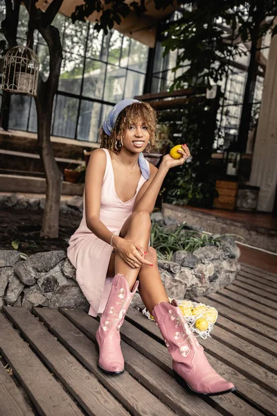 Full length of smiling young african american woman in boots and summer outfit holding fresh lemon while sitting near mesh bag in blurred orangery, chic woman in tropical garden, summer concept — Stock Photo