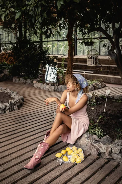 Young and trendy african american woman in headscarf, boots and summer dress holding fresh lemon and sitting near mesh bag on floor in blurred orangery, chic woman in tropical garden — Stock Photo