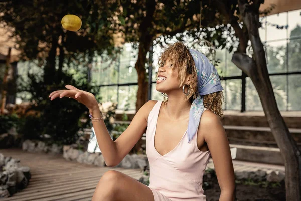 Joven mujer afroamericana alegre con frenos que usan pañuelo para la cabeza y vestido de verano mientras lanza limón maduro y se sienta en el centro del jardín borroso, mujer elegante en el jardín tropical, concepto de verano - foto de stock