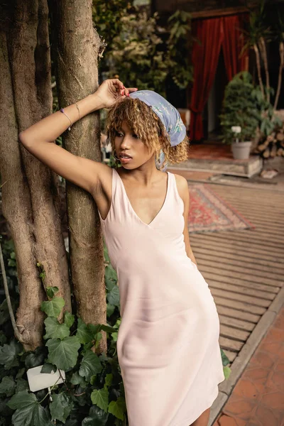 Mujer afroamericana joven de moda en pañuelo para la cabeza y vestido de verano de pie cerca de los árboles en el jardín interior borroso en el fondo, mujer elegante con plantas tropicales en el telón de fondo, concepto de verano - foto de stock