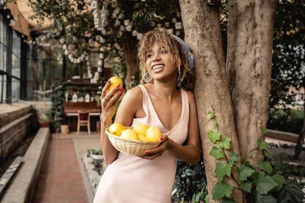 Pleased young african american woman with braces wearing summer outfit and holding ripe lemons and basket while standing near trees in orangery, fashion-forward lady in harmony with tropical flora — Stock Photo