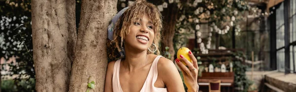 Молодая и модная африканская американка в брекетах и летнем наряде держит спелый лимон и стоит рядом с деревьями в центре сада, модная леди в гармонии с тропической флорой, баннер — стоковое фото