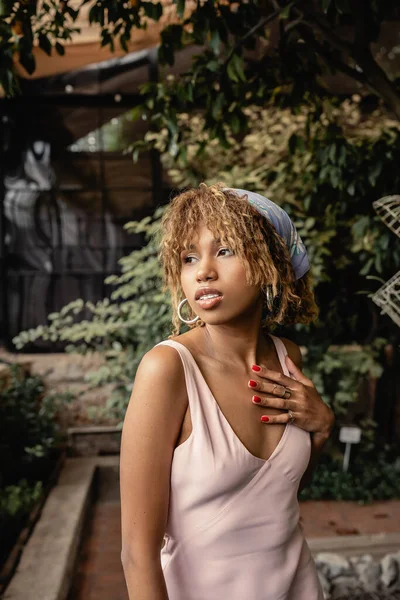 Portrait of stylish young african american woman in headscarf and summer dress touching chest and standing in blurred indoor garden, woman in summer outfit posing near lush tropical plants — Stock Photo