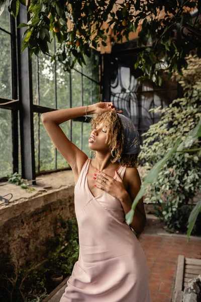 Relajada joven afroamericana mujer en traje de verano y pañuelo en la cabeza tocando el cuello mientras está de pie cerca de las plantas en el jardín interior, mujer en traje de verano posando cerca de exuberantes plantas tropicales - foto de stock