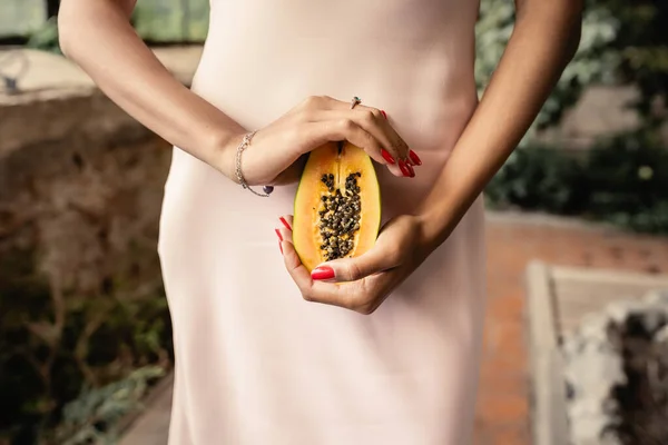 Cropped view of young african american woman in summer dress holding cut and fresh papaya while standing in blurred garden center, woman in summer outfit posing near lush tropical plants — Stock Photo