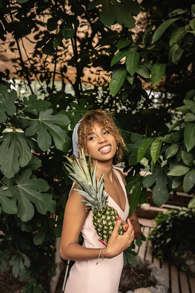 Sorrindo jovem mulher afro-americana com aparelho vestindo roupa de verão e segurando abacaxi e de pé perto de plantas em orangery, mulher em roupa de verão posando perto de plantas tropicais exuberantes — Fotografia de Stock