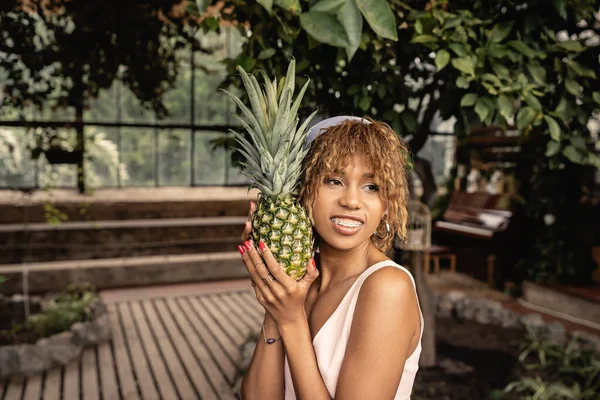 Femme afro-américaine agréable avec bretelles et tenue d'été tenant l'ananas frais et debout dans le centre de jardin, femme élégante portant une tenue d'été entourée de feuillage tropical — Photo de stock