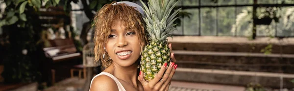 Smiling young african american woman with braces wearing headscarf and holding pineapple and standing in indoor garden, stylish woman wearing summer outfit surrounded by tropical foliage, banner — Stock Photo