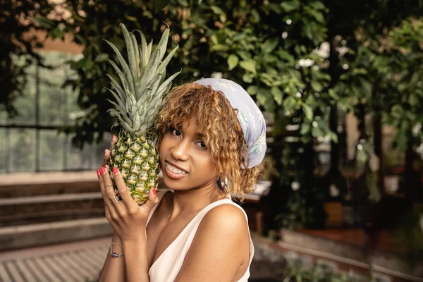 Lächelnde junge afrikanisch-amerikanische Frau mit Hosenträgern und Kopftuch, die frische Ananas hält und in verschwommener Orangerie in die Kamera blickt, stilvolle Frau im Sommeroutfit, umgeben von tropischem Laub — Stockfoto