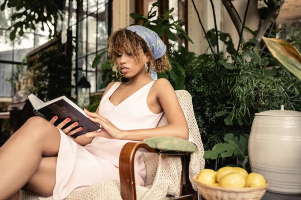 Mujer afroamericana joven de moda en traje de verano leyendo libro mientras está sentada cerca de limones borrosos en el jardín interior, mujer con traje de verano rodeada de follaje tropical, concepto de verano - foto de stock