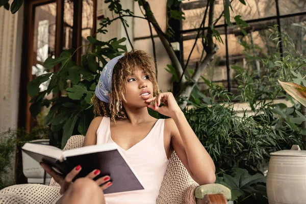 Молодая и уверенная в себе африканская женщина в летнем наряде держит книгу и сидит на кресле в размытом внутреннем саду, стильная женщина в летнем наряде в окружении тропической листвы — стоковое фото