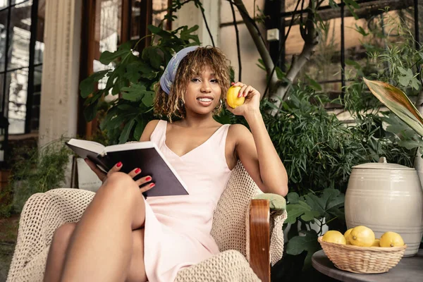 Smiling and stylish african american woman holding lemon and bool while looking at camera and sitting on armchair in garden center, woman in comfortable and trendy summer outfit — Stock Photo