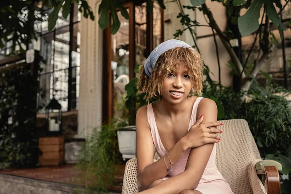 Joyeux jeune femme afro-américaine avec bretelles et robe d'été regardant la caméra tout en étant assis sur un fauteuil dans le jardin intérieur moderne, femme dans une tenue d'été confortable et à la mode — Photo de stock