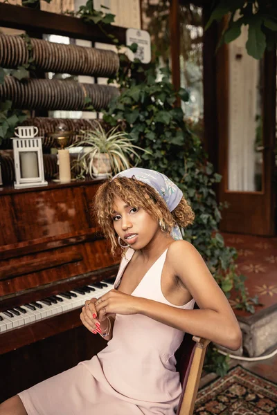 Moderne junge afrikanisch-amerikanische Frau im Sommerkleid, die in die Kamera blickt, während sie neben Klavier und verschwommenen Pflanzen in der Orangerie sitzt, Frau im bequemen und trendigen Sommeroutfit — Stockfoto
