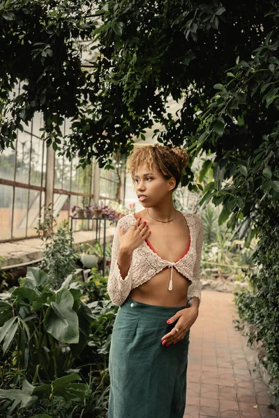 Trendy giovane donna afroamericana in maglia top e gonna in posa e in piedi sotto le piante in giardino coperto offuscata, donna alla moda con piante tropicali sullo sfondo — Foto stock