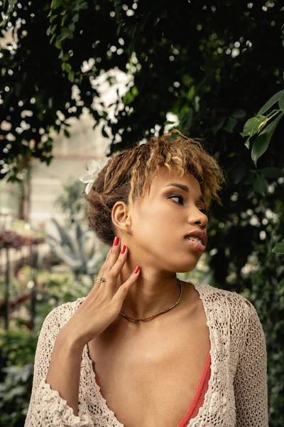 Porträt einer jungen afrikanisch-amerikanischen Frau im Stricktop, die den Hals berührt und neben grünen Pflanzen im Gartencenter steht, modebewusste Frau mit tropischen Pflanzen im Hintergrund, Sommerkonzept — Stockfoto