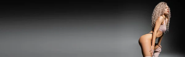 Modello femminile sexy con capelli biondi cenere ondulati in posa seducente e distogliendo lo sguardo su sfondo nero e grigio, biancheria intima di pizzo, sessualità e moda, banner con spazio copia — Foto stock