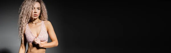 Donna affascinante e sognante con capelli biondi cenere ondulati e corpo snello in posa in reggiseno di pizzo e guardando lontano su sfondo nero e grigio, individualità sensuale, look sexy, banner con spazio copia — Foto stock