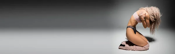 Piena lunghezza della donna sexy con corpo snello inginocchiato e abbracciandosi su sfondo grigio, biancheria intima di pizzo, stivali alti, viso oscurato con capelli biondi cenere ondulati, banner con spazio copia, vista laterale — Foto stock