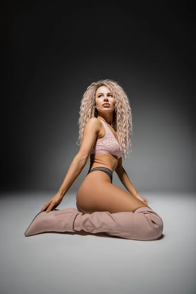 Повна довжина чарівної жінки з тонким тілом і пофарбованим попелом блондинка сидить і дивиться на чорно-сірий фон, мереживна білизна, високі чоботи, сексуальна мода і жіночність — стокове фото