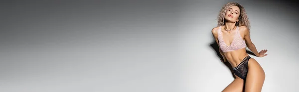Sexy Frau mit geschlossenen Augen und welligen aschblonden Haaren sitzt in aufreizender Pose auf grauem Hintergrund, Spitzenunterwäsche, durchtrainierter Körper, Weiblichkeit und Stil, Hochblick, Banner mit Kopierraum — Stockfoto