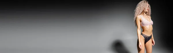Mulher atraente com corpo tonificado e cabelo loiro cinza tingido em pé no fundo preto e cinza e olhando para longe, roupa interior de renda, tendência de moda sexy, banner com espaço de cópia — Fotografia de Stock