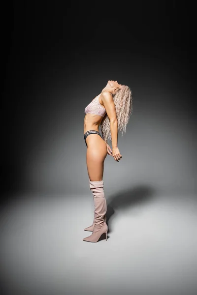 Sexuality and fashion, modern self-expression, full length of female model with toned body and wavy ash blonde hair posing in lace underwear and high stylish boots on grey background, side view — Stock Photo