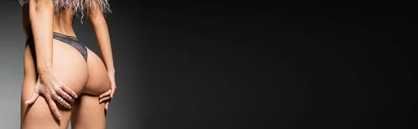 Sexualität und Mode, abgeschnittene Ansicht einer provokanten Frau mit durchtrainiertem Körper, die sexy Gesäß berührt, während sie in Spitzenhöschen auf schwarzem und grauem Hintergrund posiert, Banner mit Kopierraum — Stockfoto
