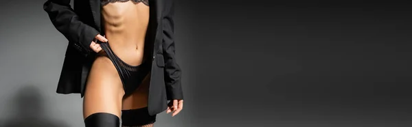 Vista recortada de la mujer glamour con el cuerpo tonificado ajustando las bragas mientras posan en chaqueta negro y medias sobre fondo gris, sexualidad y moda, pancarta con espacio para copiar - foto de stock