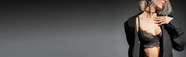 Vista parcial de la mujer sensual con la ceniza ondulada pelo rubio posando en sujetador de encaje y blazer negro y tocando el pecho sobre fondo gris, aspecto sexy, glamour y feminidad, pancarta con espacio para copiar - foto de stock
