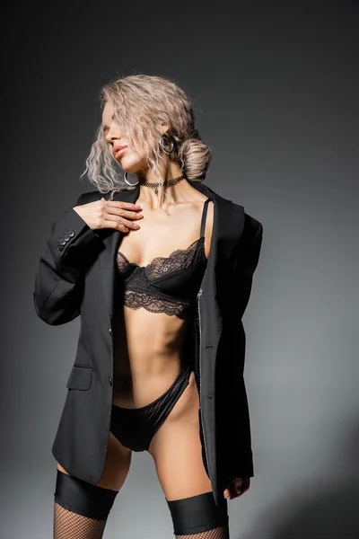 Femme avant-gardiste et sexy en blazer noir, culotte et soutien-gorge en dentelle posant sur fond gris, cheveux blonds cendrés teints et ondulés, représentation de lingerie élégante sur un corps mince — Photo de stock