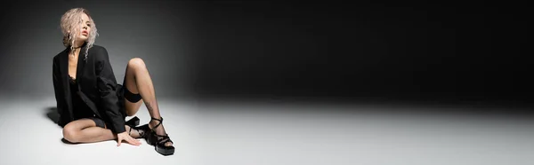 Volle Länge der modischen und verführerischen Frau mit aschblonden Haaren und schlankem Körper sitzt im Blazer, Strümpfe und Sandalen mit Absatz auf schwarzem und grauem Hintergrund, sexy Stil, Banner mit Kopierraum — Stockfoto