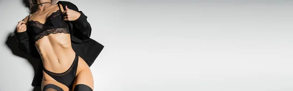Confianza y auto-expresión, vista parcial de la mujer sensual con el cuerpo tonificado en blazer negro, medias, bragas y sujetador de encaje sobre fondo gris, pancarta con espacio para copiar, vista superior - foto de stock