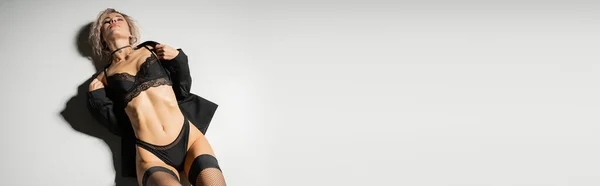 Vista superior de mujer atractiva con cuerpo tonificado y pelo rubio de ceniza ondulada, en lencería elegante, chaqueta negra y medias que ponen sobre fondo gris, fotografía de moda sexy, pancarta con espacio para copiar - foto de stock