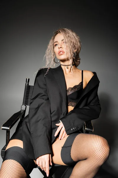 Elegante modella donna in blazer nero, lingerie e calze a rete seduta sulla sedia e distogliendo lo sguardo su sfondo grigio, capelli biondi cenere ondulati, sguardo espressivo, glamour, stile di vita sexy — Foto stock