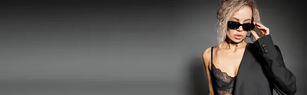 Mulher fashion-forward com cabelo loiro cinza tingido, usando sutiã de renda e blazer, ajustando óculos escuros e em pé sobre fundo cinza, olhar sexy, confiança, banner com espaço de cópia — Fotografia de Stock